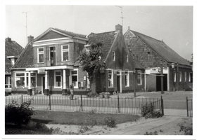 Zwart-witfoto van De Postwagen met hekwerk en de buurpanden aan de Hoofdstraat