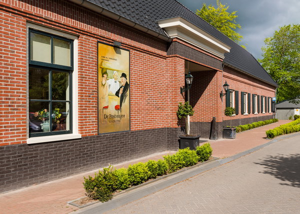 Zijgevel van De Postwagen met nostalgische reclameposter en groene heggen